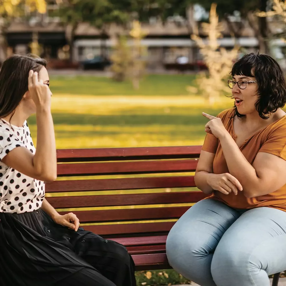 Two women sit on a park bench one woman is signing to the other